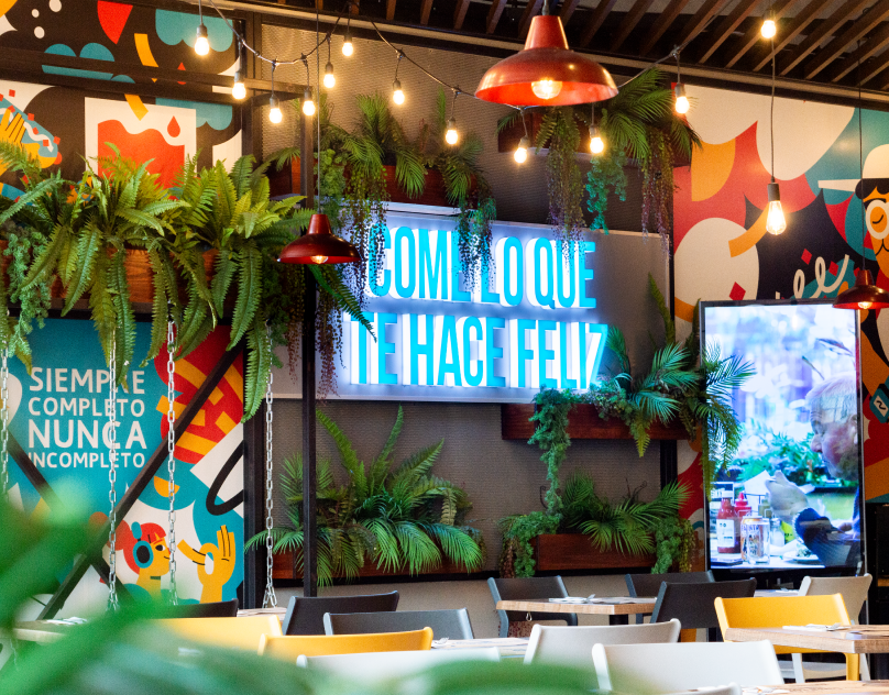 Wide shot of a dining room with a neon sign that reads Come lo que te hace feliz over mural art.