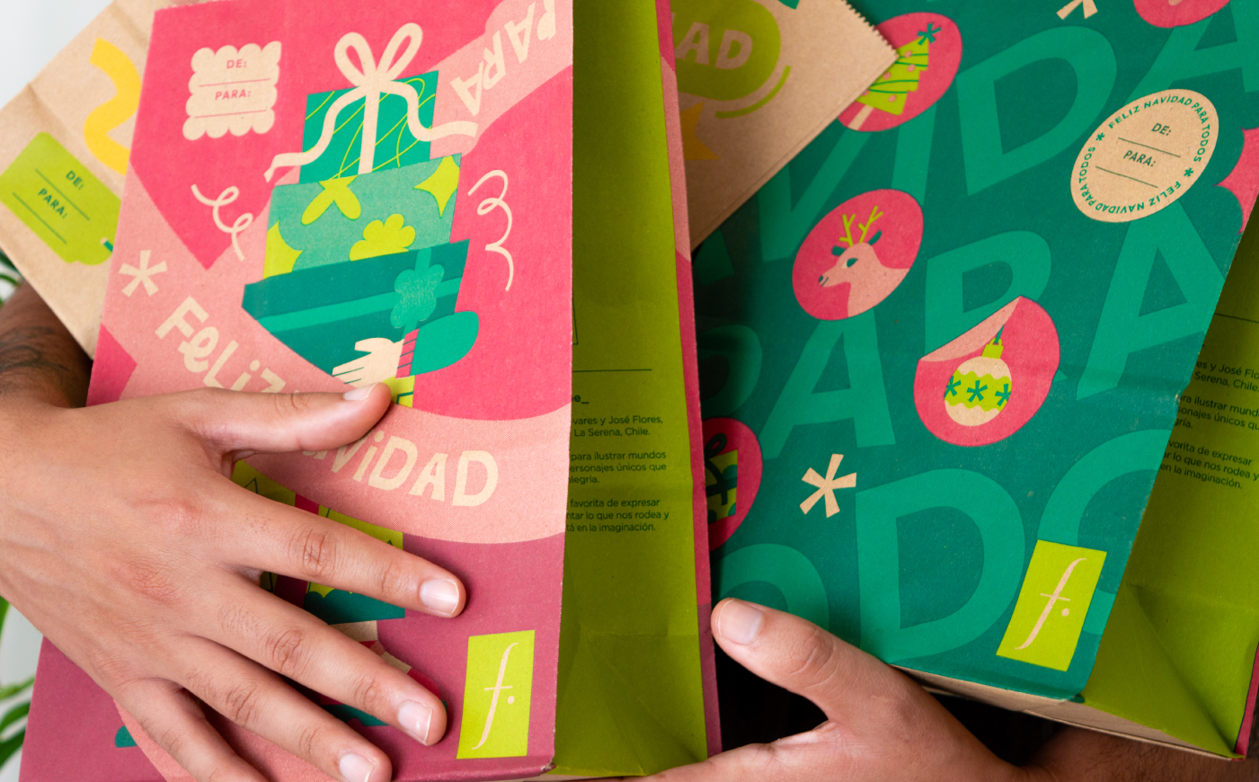 Person holding two illustrated paper gift bags with vibrant Christmas character designs and festive typography.